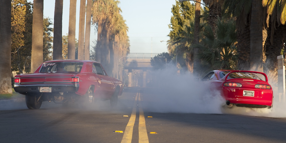 1967-chevy-chevelle-burnout-with-mk3-toyota-supra Two way road with red 1967 Chevy Chevelle and bright red MKR Toyota Supra smoking the tires with a huge burnout on either side of the double yellow line in the middle of the road